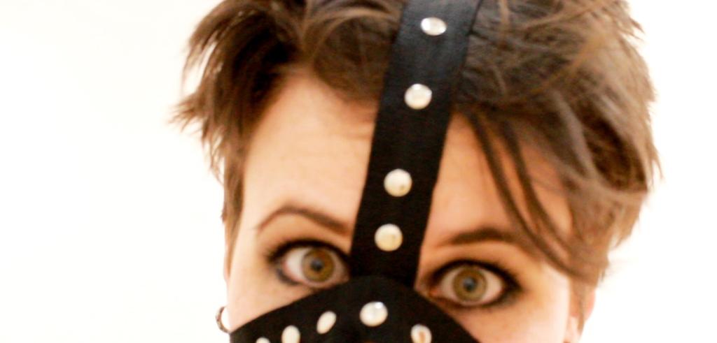 Photo of the top half of the artist's face, covered in a muzzle-like piece of fabric. The artist has pale skin, brown hair, and wears dark black eye makeup. 