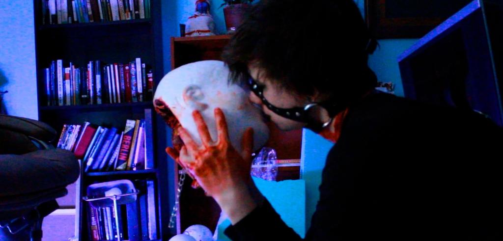 Color photo of the artist wearing a muzzle-like harness around their face, holding a life-size head sculpture in bloodied hands in front of blue-toned bookshelves. 