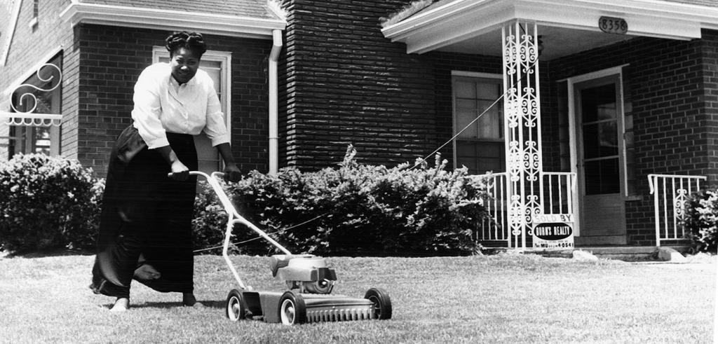 An African American woman in a '50s suburb setting mowing her home's lawn with a push mower, while smiling at the camera. 