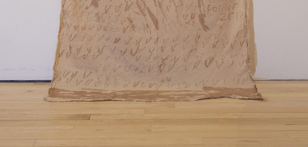 Tan tapestry with darker tan abstract shapes on it, hanging partially against a white wall and draped on wooden floor.