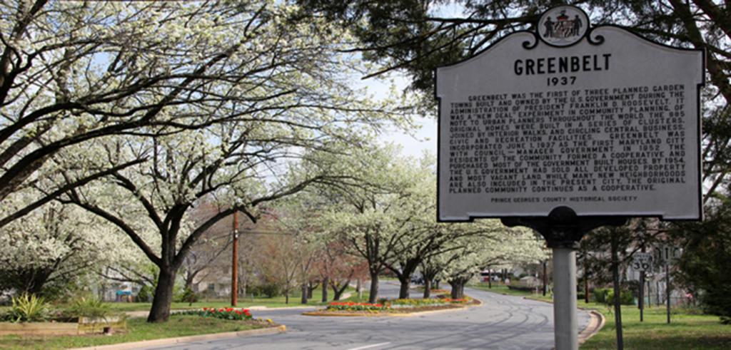 Historic marker sign for Greenbelt, Maryland, with a street behind it and trees in bloom