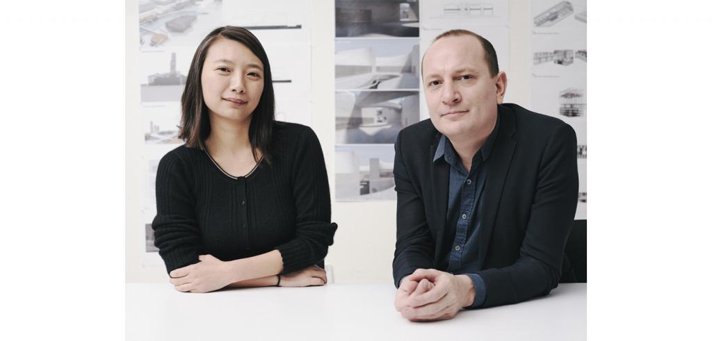 A man and a woman, each in black tops looking at the camera, with a wall of architectural renderings behind.