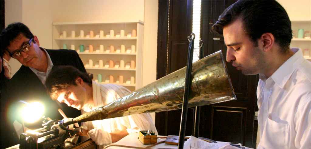A person with short black hair blows into a conical brass tube with a light at the end of it. 