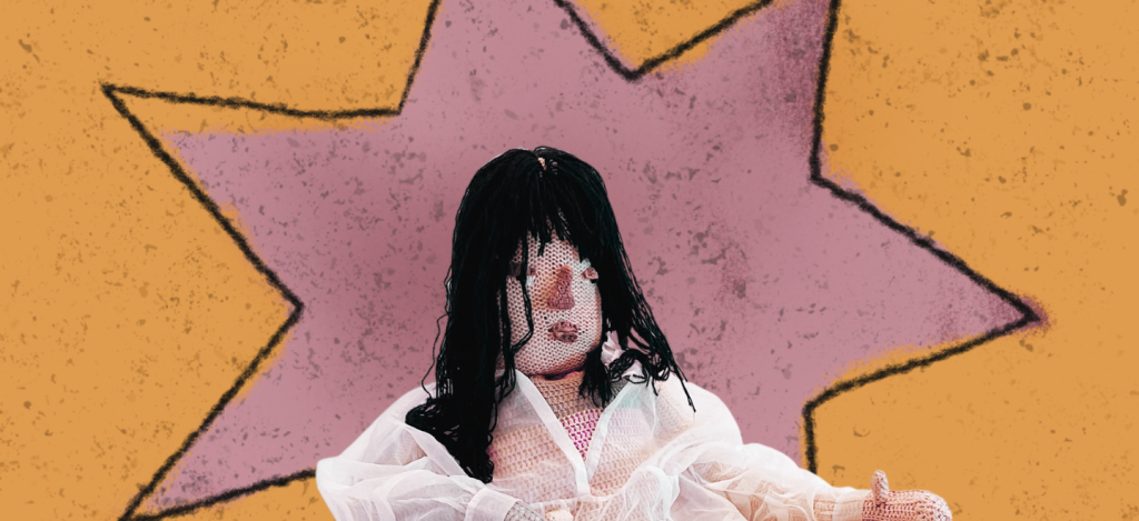 Color photo of a crocheted human doll with black hair wearing a chiffon blouse in front of a pink and orange abstract background. 