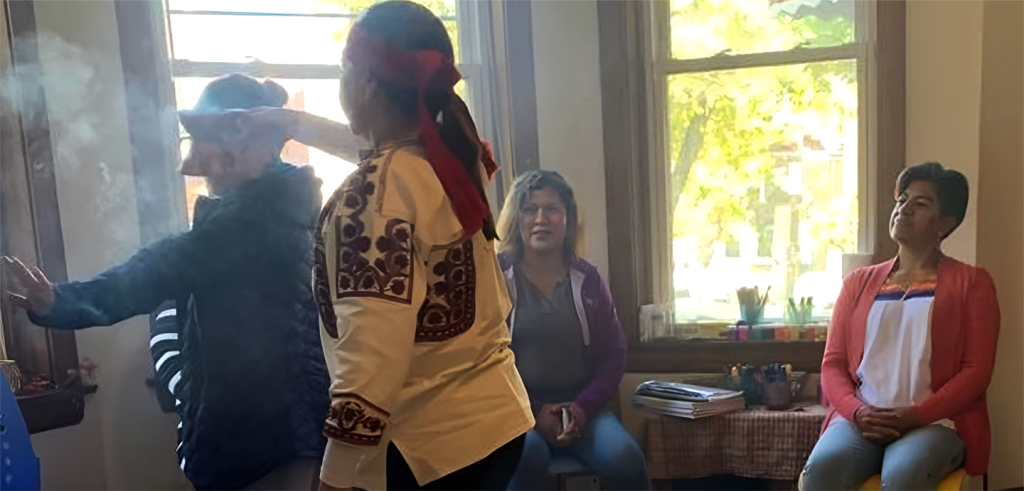 Group inside a room with large windows. One person wearing a red head wrap is using smoke next to another person. Others are sitting in chairs watching.