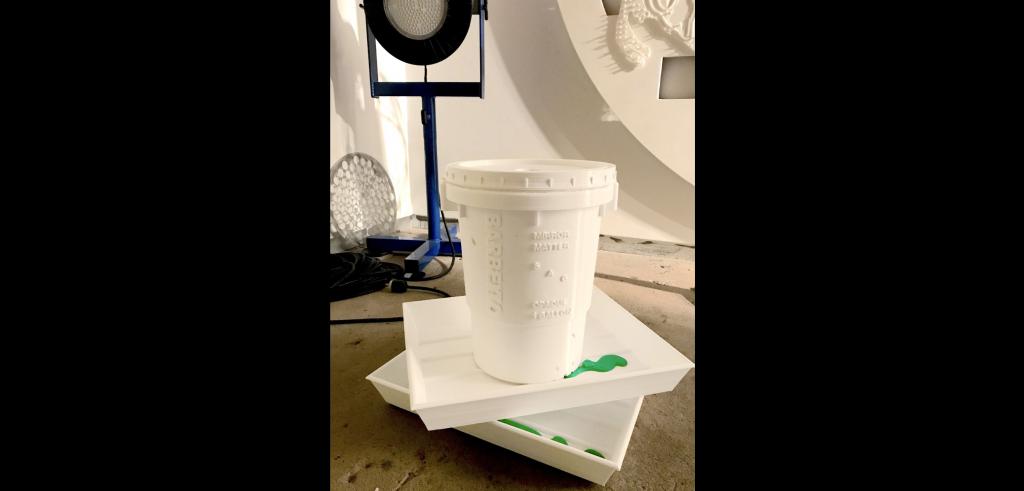 White bucket on two square white trays with a little green paint on them, on a cement floor with a white background and blue colored light.