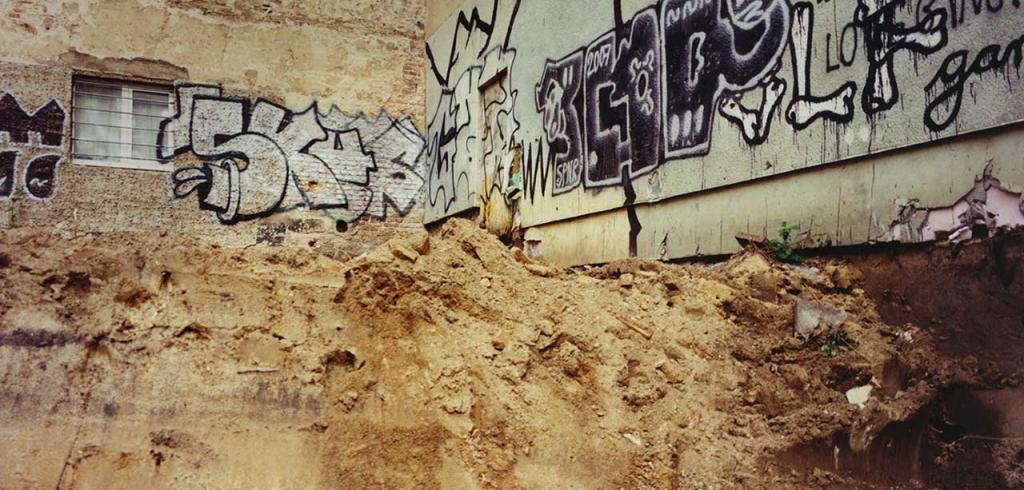 A concrete wall with crumbling pieces, black and white graffiti, and a large dirt pile in front of it.