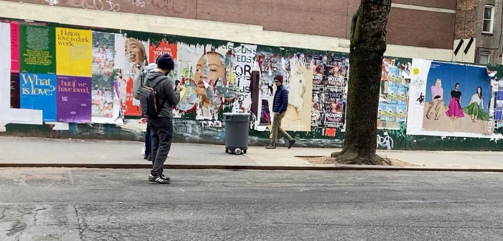 Horizontal color photo of three people on a concrete city block, looking at a trashcan on motorized wheels. Behind the trash can is a wall pasted with torn advertisements and posters. 