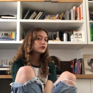 A person sitting with their legs crossed with ripped jeans and a white bookshelf in the background.