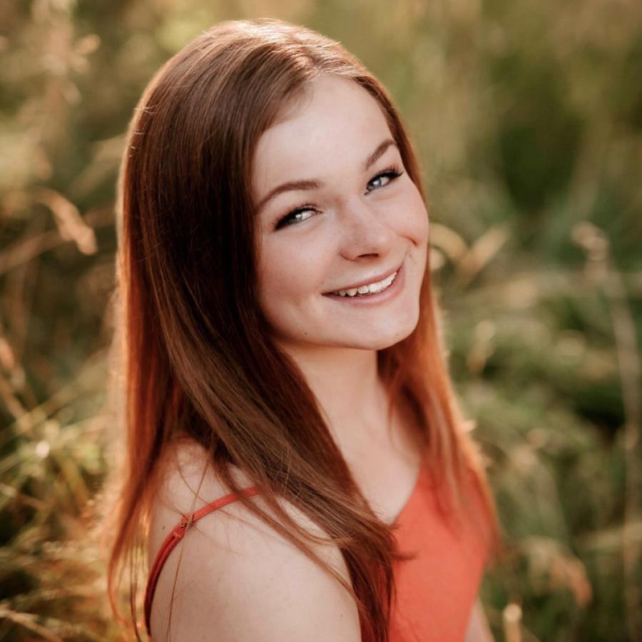 A headshot of a person smiling with green and yellow foliage in the background.