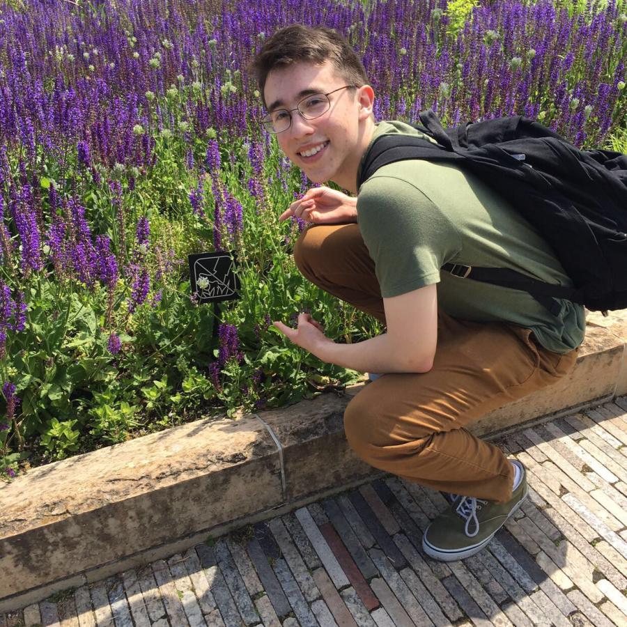 student in green shirt, khakis, black backpack on a wood bridge with purple flowers