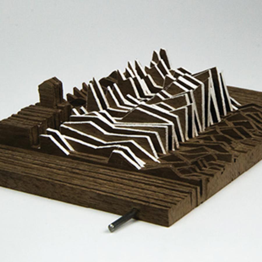 Photograph of model made from vertical strips of chipboard stacked together to form a topological mass, strung together by two metal rods, with a portion in the center showing more extreme angular profiles having strips of white paper bridging between the chipboard.