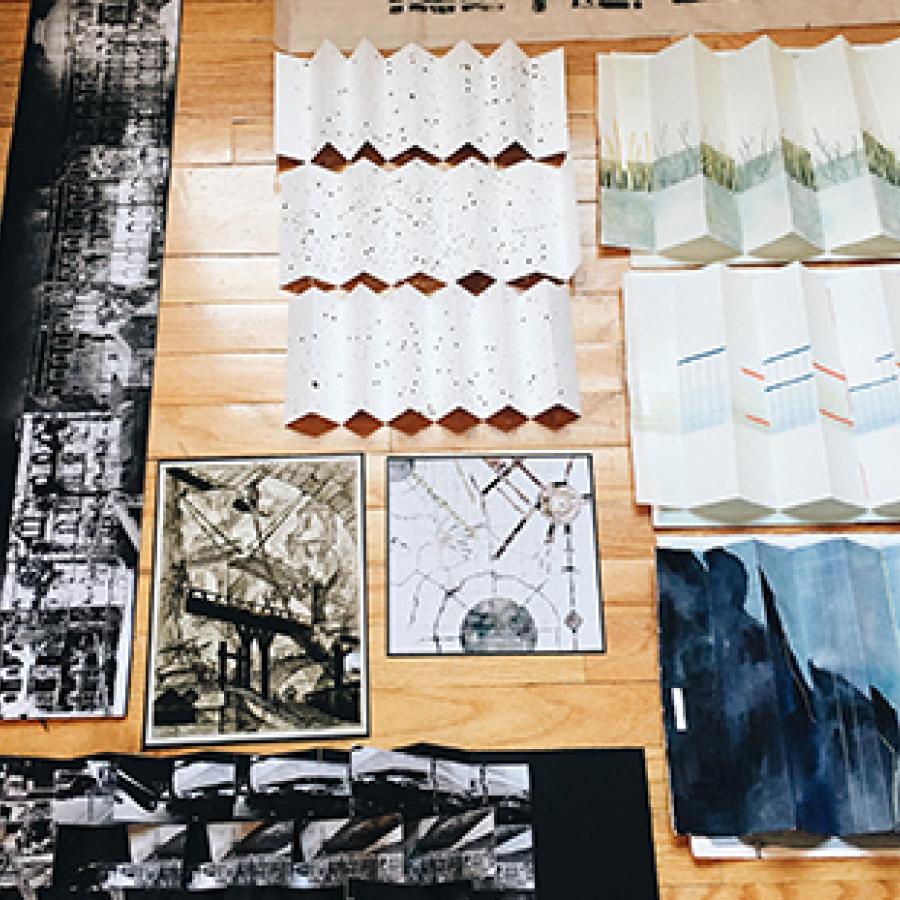 Photograph of set of drawings and images in different mediums, some folded, laid out on a wooden floor.