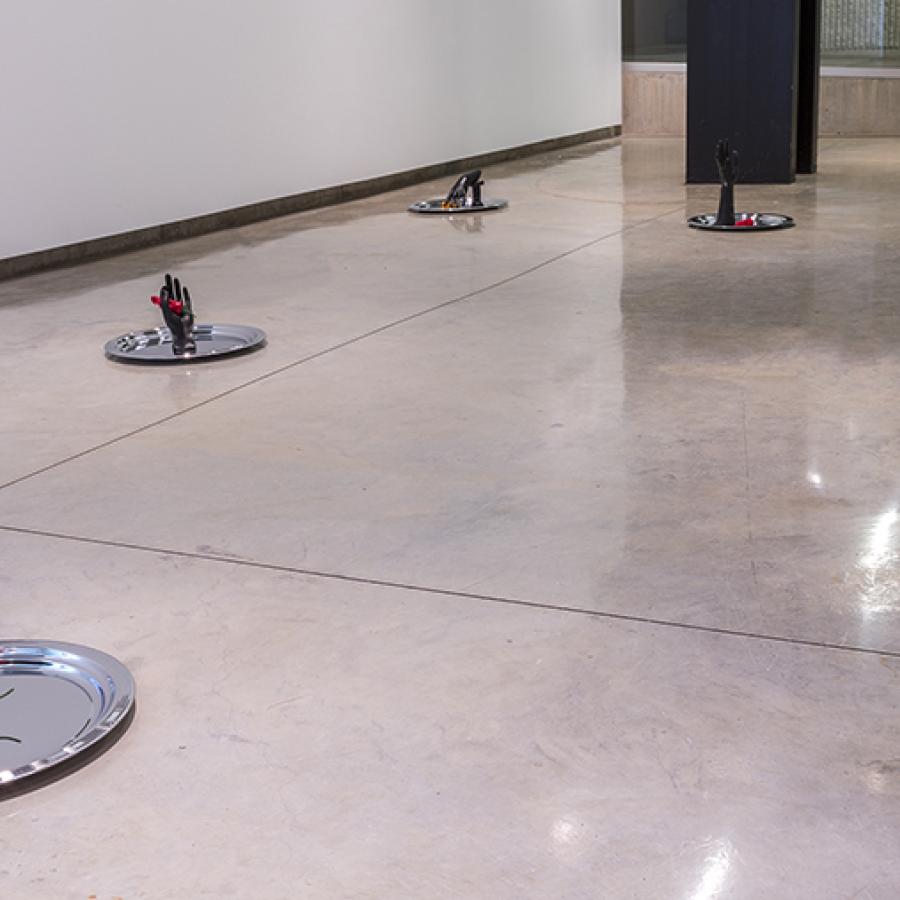Six silver platters on a cement floor with black hands and feet on each one along with a red or orange colored pepper.