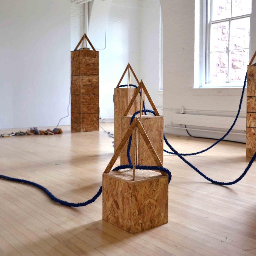 Wooden obelisk statues of various size with blue rope connecting them together in a white walled gallery with a light wood floor and long windows which bring sunlight in. 