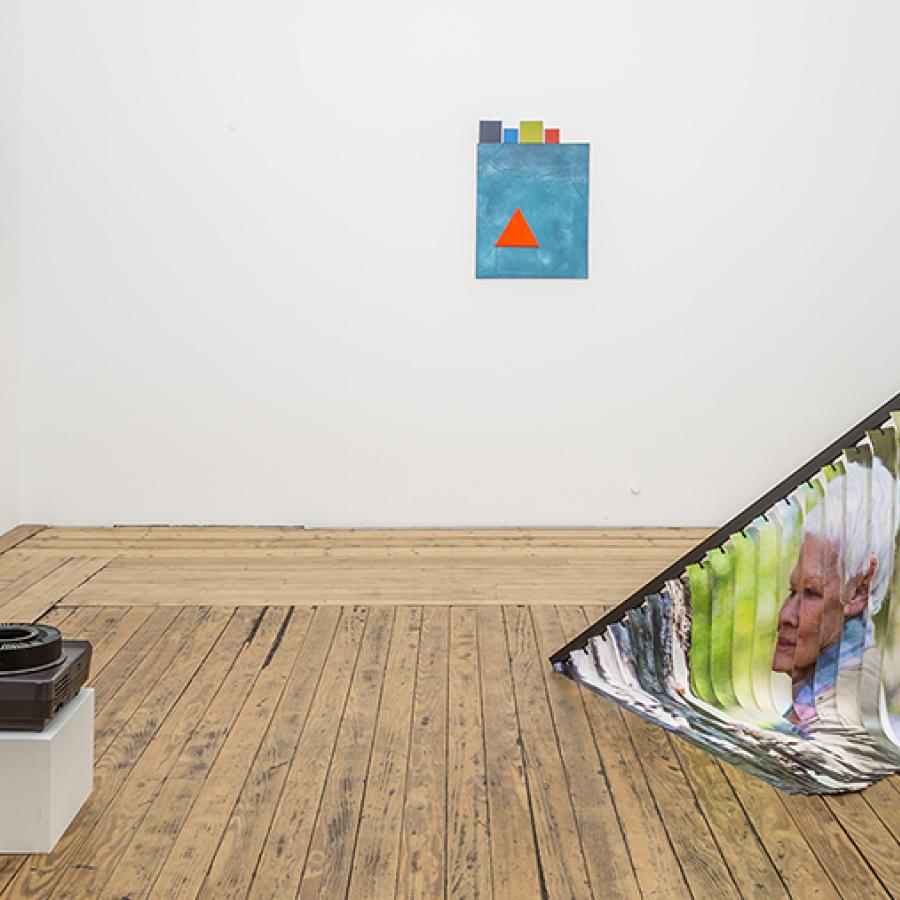 Small slide show projector, three paintings on the wall in various hues of orange and blue with Popsicle sticks and a black pedestal with vertical blinds draped towards the floor with Judi Dench's face.