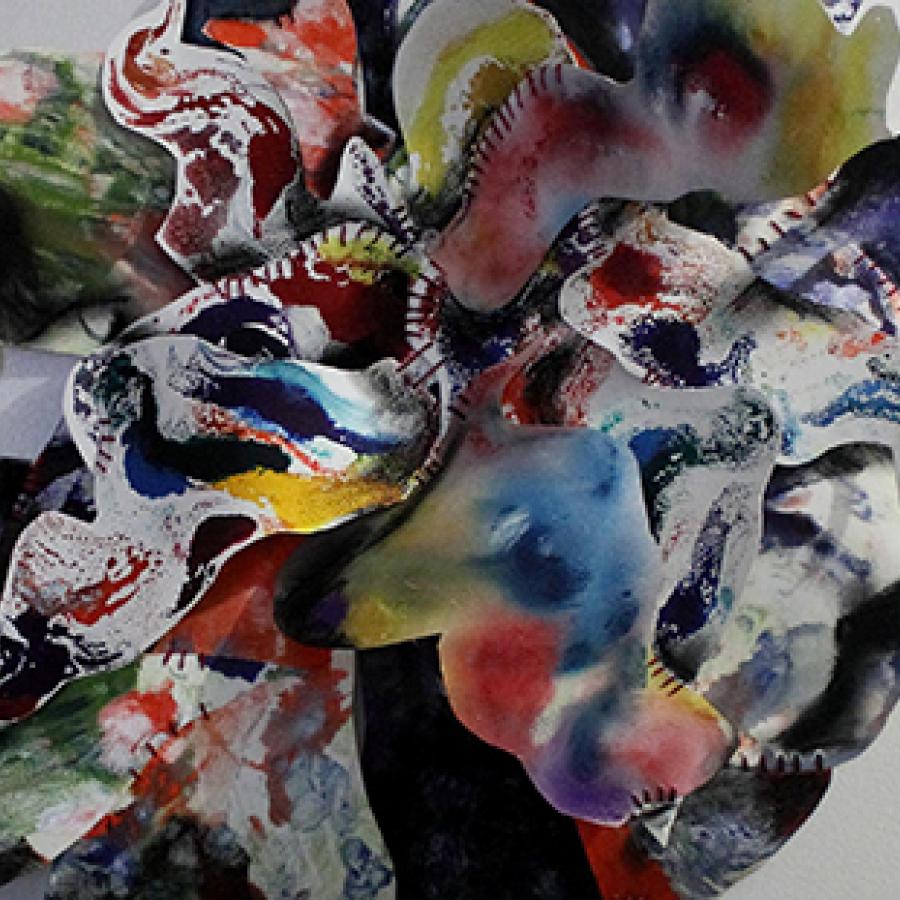 Color photo of an abstract flower-like sculpture made of colorful watercolor on white paper, hung on a white wall. 