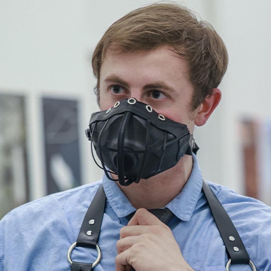 Blonde white man dressed in a blue-collar t-shirt wearing a black leather harness and with metal rings and a leather-looking maskwith a speaker in place of its mouth.
