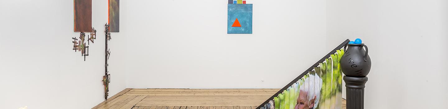 Small slide show projector, three paintings on the wall in various hues of orange and blue with Popsicle sticks and a black pedestal with vertical blinds draped towards the floor with Judi Dench's face.