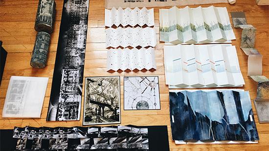 Photograph of set of drawings and images in different mediums, some folded, laid out on a wooden floor.