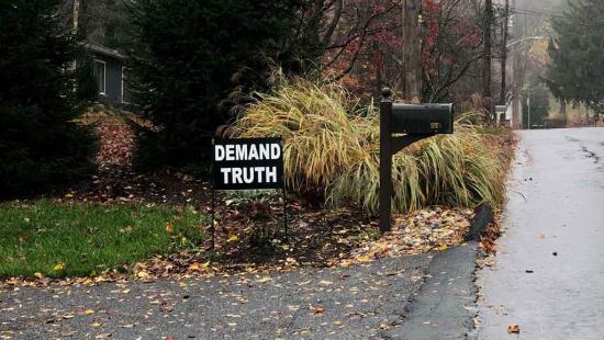 Black and white sign depicting the words DEMAND TRUTH next to a mailbox and on the corner of someone's lawn.