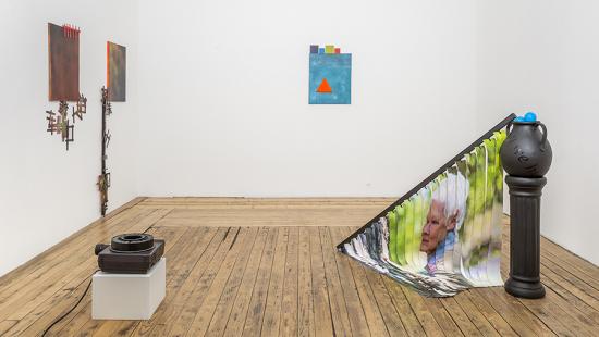 Small slide show projector, three paintings on the wall in various hues of orange and blue with Popsicle sticks and a black pedestal with vertical blinds draped towards the floor with Judi Dench's face.