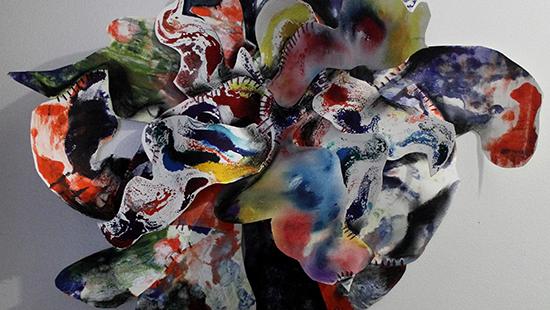 Color photo of an abstract flower-like sculpture made of colorful watercolor on white paper, hung on a white wall. 