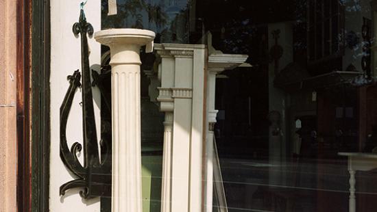 Color photograph of two free standing architectural columns, one inside of a storefront window (partially shaded) and the other outside of the window in the full sunlight.
