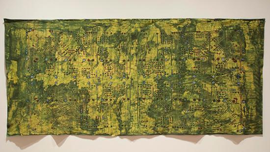 Color photograph of textile art hung on a white wall. The muslin fabric base is green and mustard colors, with black designs reminiscent of computer chips and electronic pathways painted on in black. 