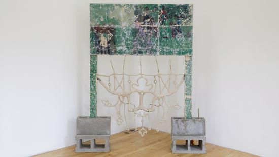 A wooden frame with a printed design in shades of green and purple, held up by concrete blocks, with an intricate white clay-like sculpture hanging from it. 