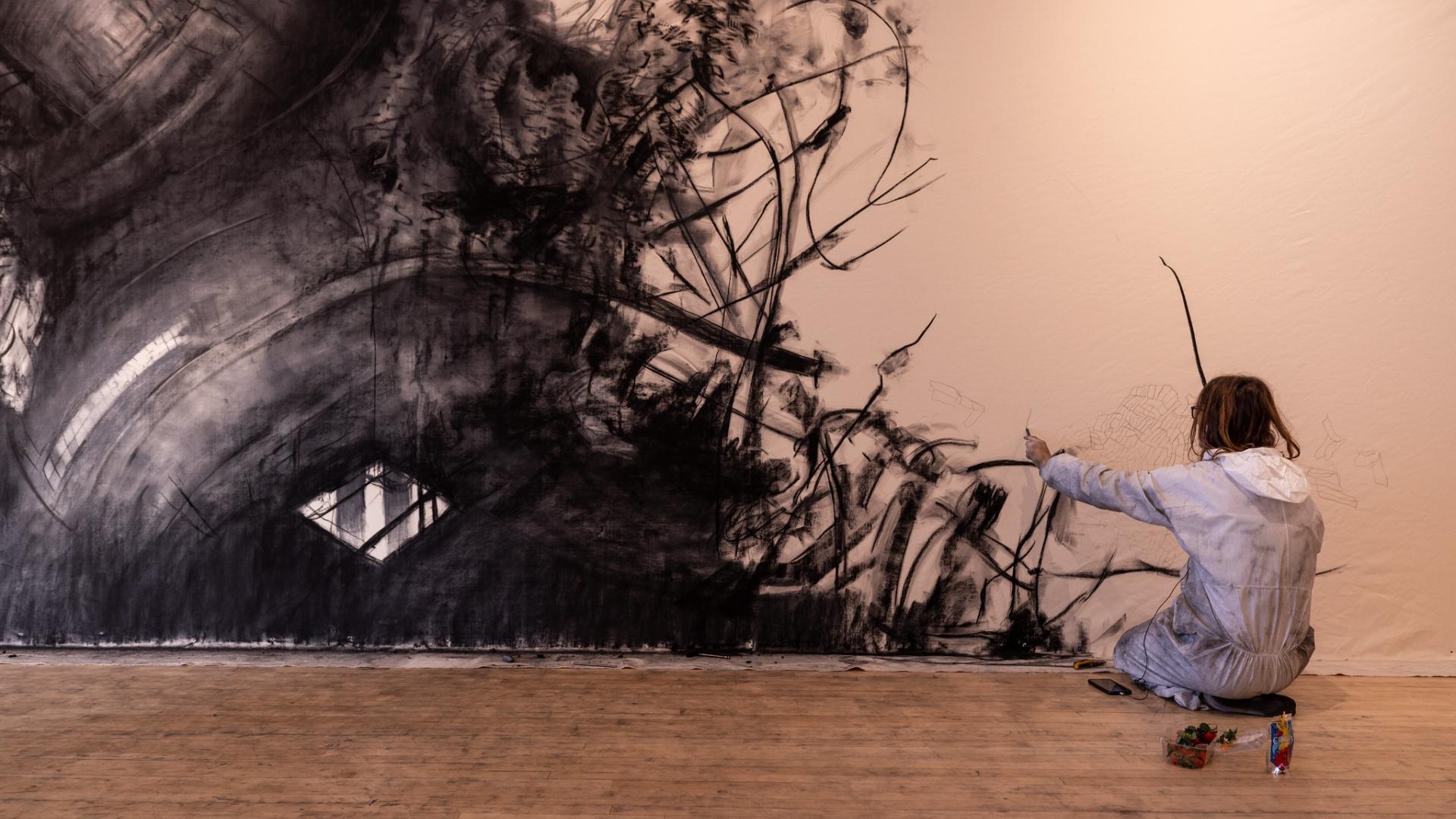 A person kneeling down observing their artwork after drawing on the wall with charcoal.
