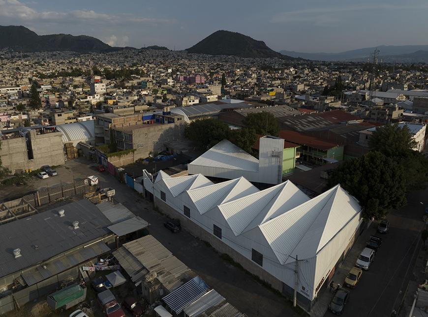 Aerial view of the Pilares Quetzalcóatl community center in Mexico City. The building has a series of interconnected white roofs with a central courtyard and is surrounded by urban development.