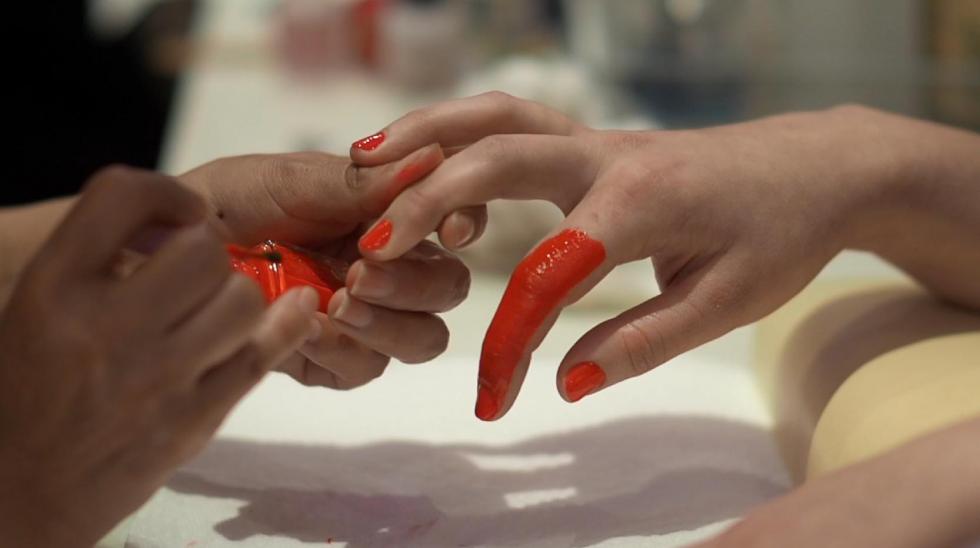 Two hands holding bright red nail polish as it paints another hand's fingernails with the index finger fully painted red.