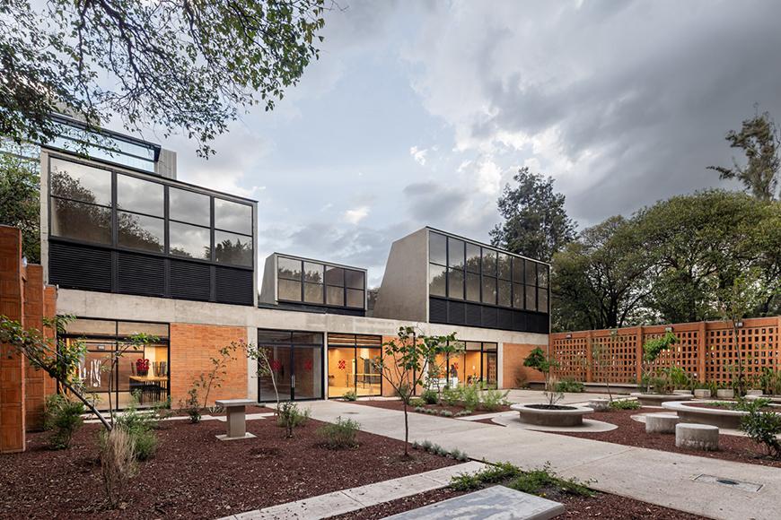 A modern community center with exposed brick walls and concrete frames. The building has a central courtyard with greenery and seating areas. Large windows offer natural light and views of the outdoors.