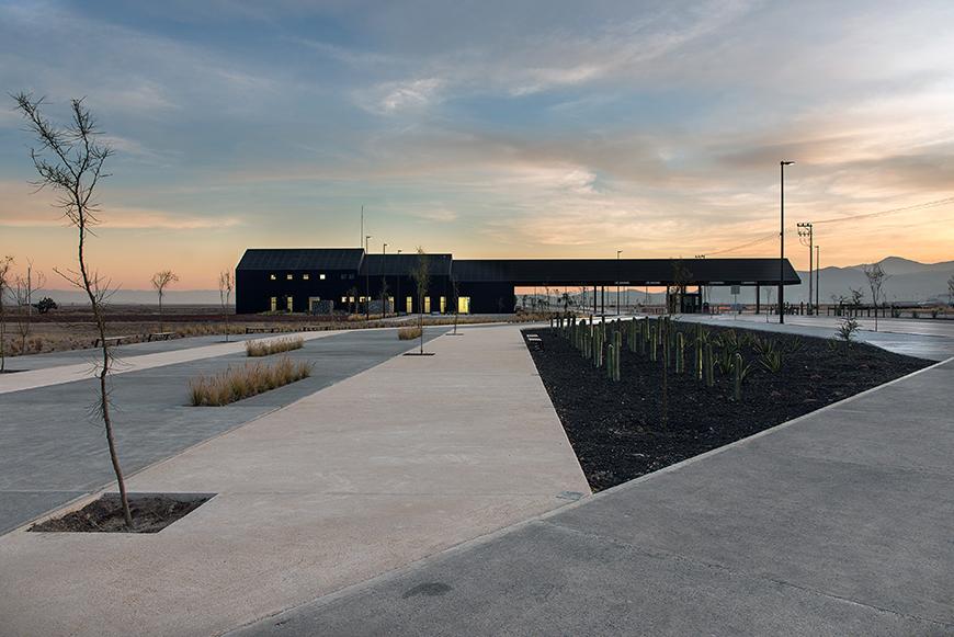 A modern, black building with a flat roof and large windows sits on a concrete plaza. The building has a covered walkway leading to its entrance. There are several small trees and shrubs planted along the plaza, and a clear blue sky with white clouds can be seen in the background.