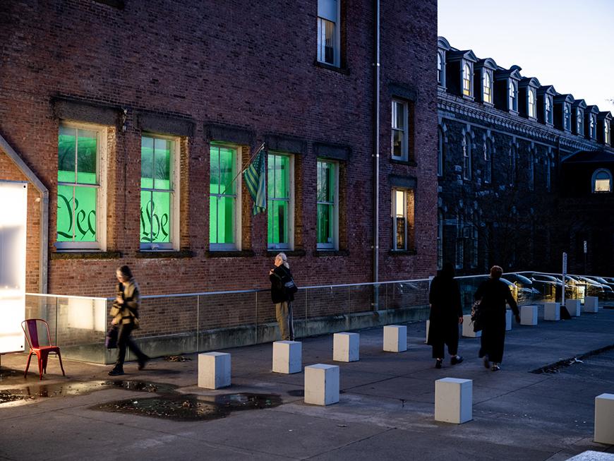 An outside view of a brick building with people looking through the windows at an exhibition with green lighting and an American flag and glimpses of the words 