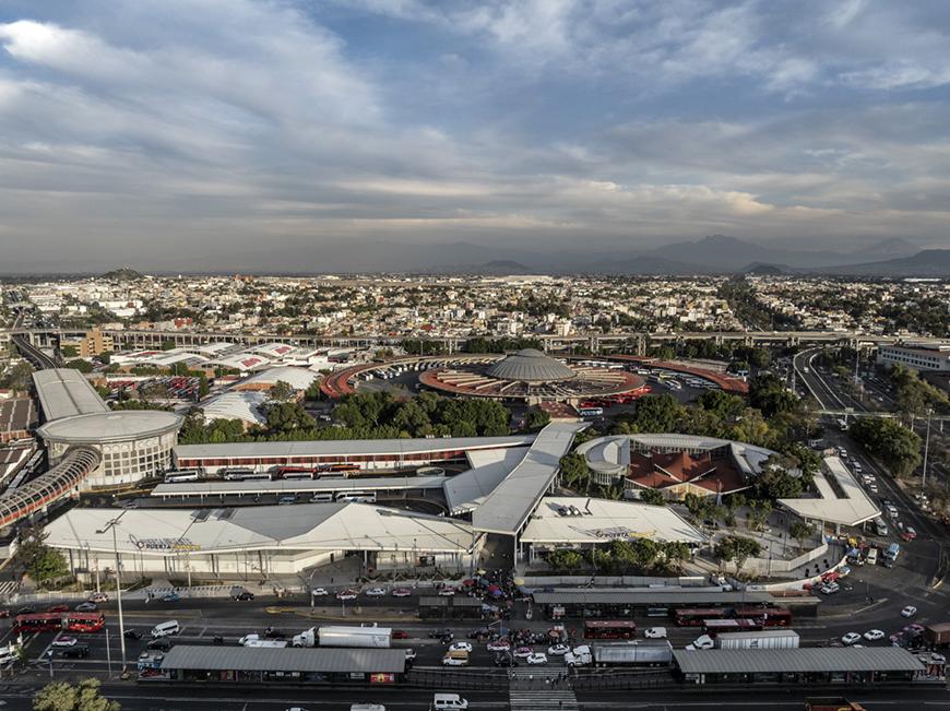 An aerial view of a large, modern bus terminal complex in a bustling city. The terminal has multiple levels, with numerous bus bays and walkways connecting the different sections. There are also green spaces and a central dome-shaped structure. The city skyline and surrounding highways are visible in the background.