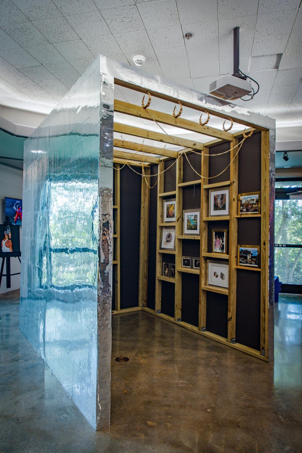 An artistic installation in a gallery featuring a reflective wall on the outside and on the interior a wooden frame with exposed beams and black walls. Framed photographs are displayed.