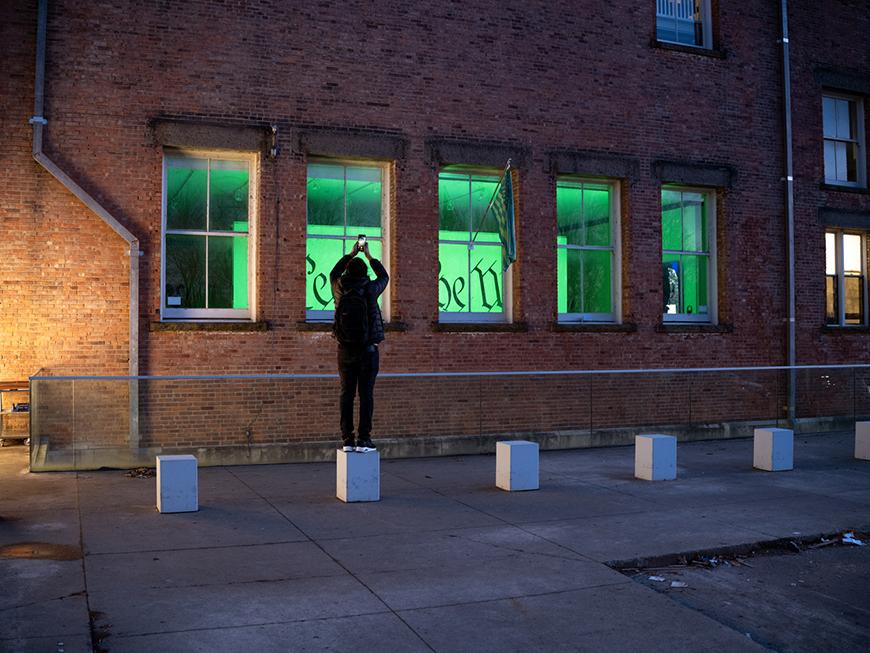 An outside view of a brick building with a person taking a photo and looking through the windows at an exhibition with green lighting and an American flag and glimpses of the words 