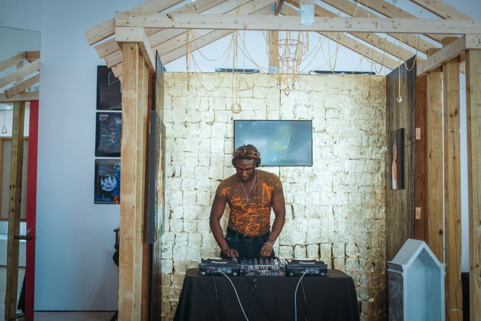 A focused DJ with headphones mixes music at a booth set against a textured gold wall inside a rustic wooden frame, surrounded by hanging chains and various artworks.