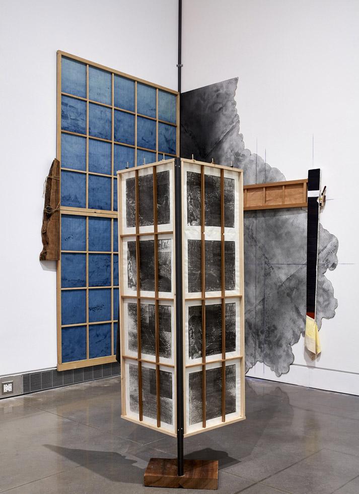 Abstract sculpture of two rectangular pieces with four black panels, with two brown lines down the middle, on a small wooden platform in front of a blue multipaneled piece and black abstract piece.