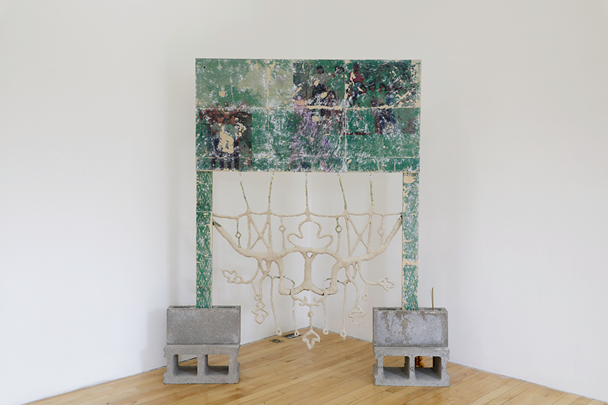 A wooden frame with a printed design in shades of green and purple, held up by concrete blocks, with an intricate white clay-like sculpture hanging from it. 