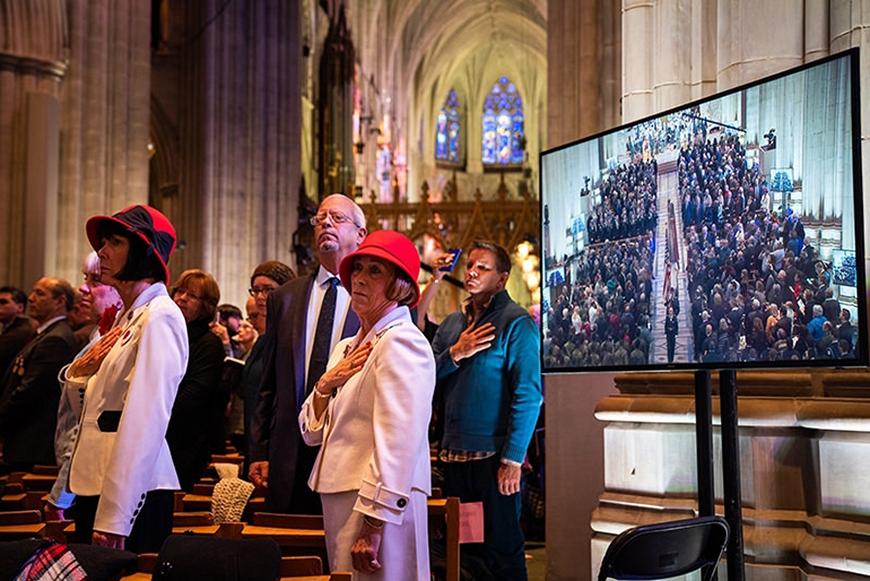 The interior of an open church space, occupied by attendees placing hands over their hearts. 