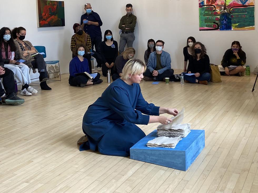 Person in a dark blue dress kneeling in front of a painted blue pedestal with two piles of cloth in front of a masked audience.