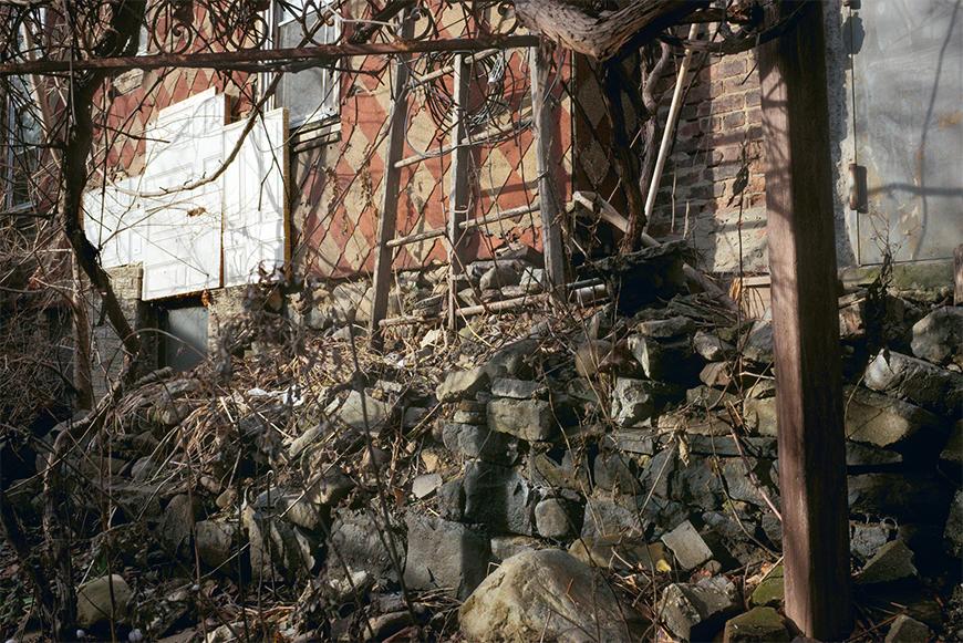 A crumbling wall with vines and weeds covering it in front of a building with a red and tan diamond pattern.