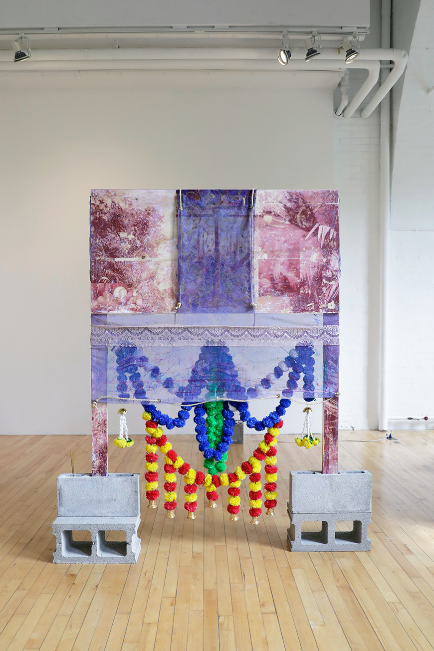 A wooden frame draped in purple and blue fabric, held up on concrete blocks, with colorful balls of blue, red and yellow balls hanging from it.