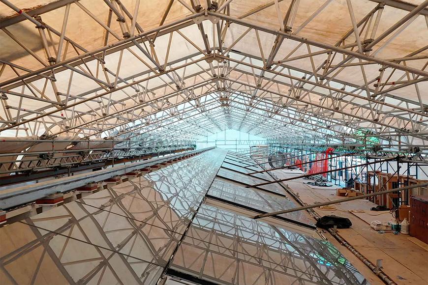  A photo of a large, open-air space with scaffolding covering the ceiling. The space appears to be a building under construction, with exposed beams and pipes visible. There is a reflective surface on the floor, possibly a glass roof or skylight.