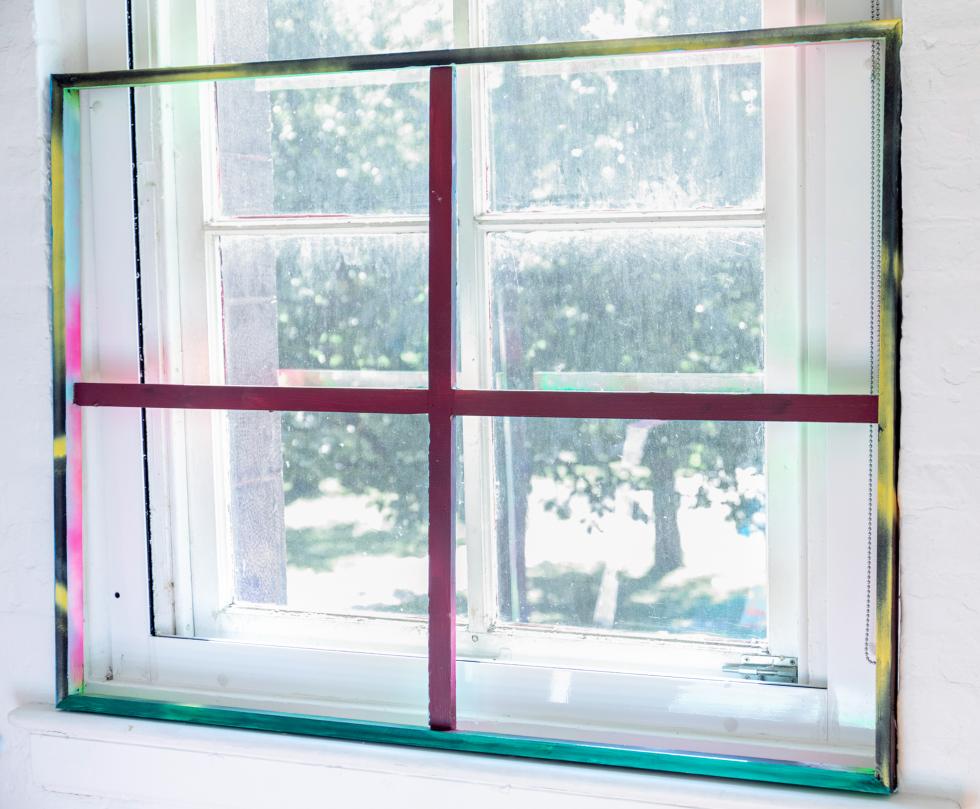 Painted window frame in pink, yellow, blue, green colors set against a white window overlooking a large tree.