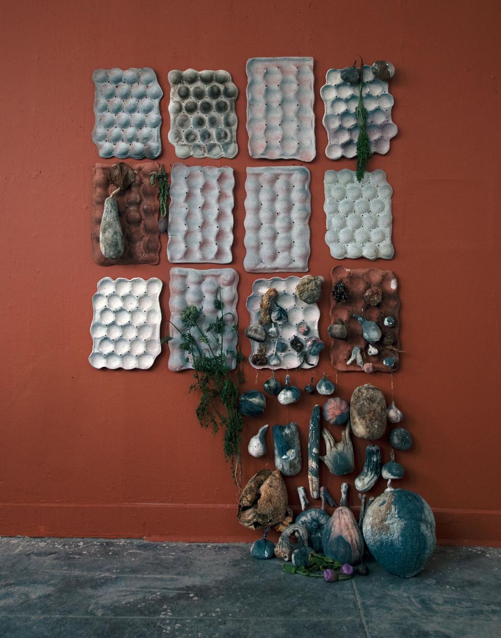 Dark red wall with 12 different colored gray egg cartons and randomly placed clay gourds hanging from them with larger clay gourds in the bottom left.