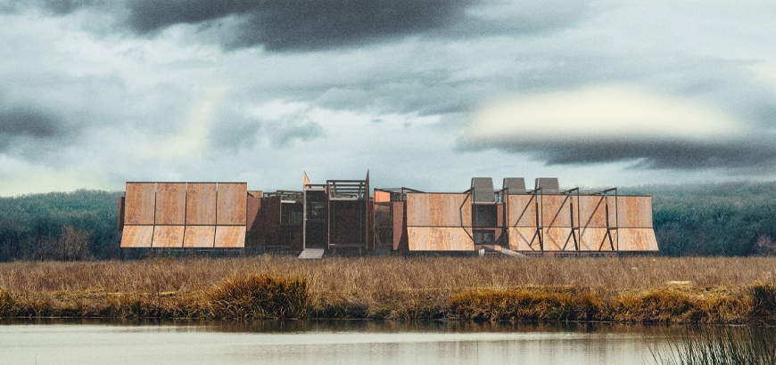 A large rusty building in an empty field with water and dark clouds in the sky.